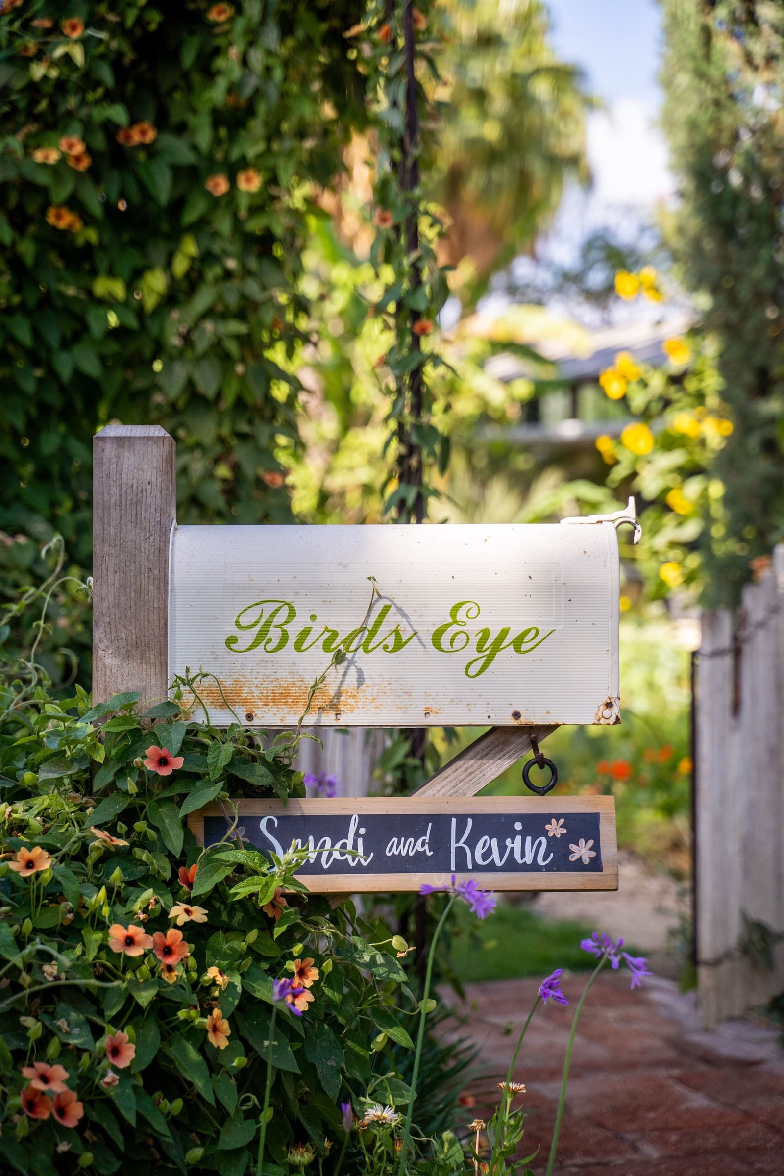 Close-up of a Flora Farms cottage mailbox with a hand-lettered name sign, surrounded by flowering plants.