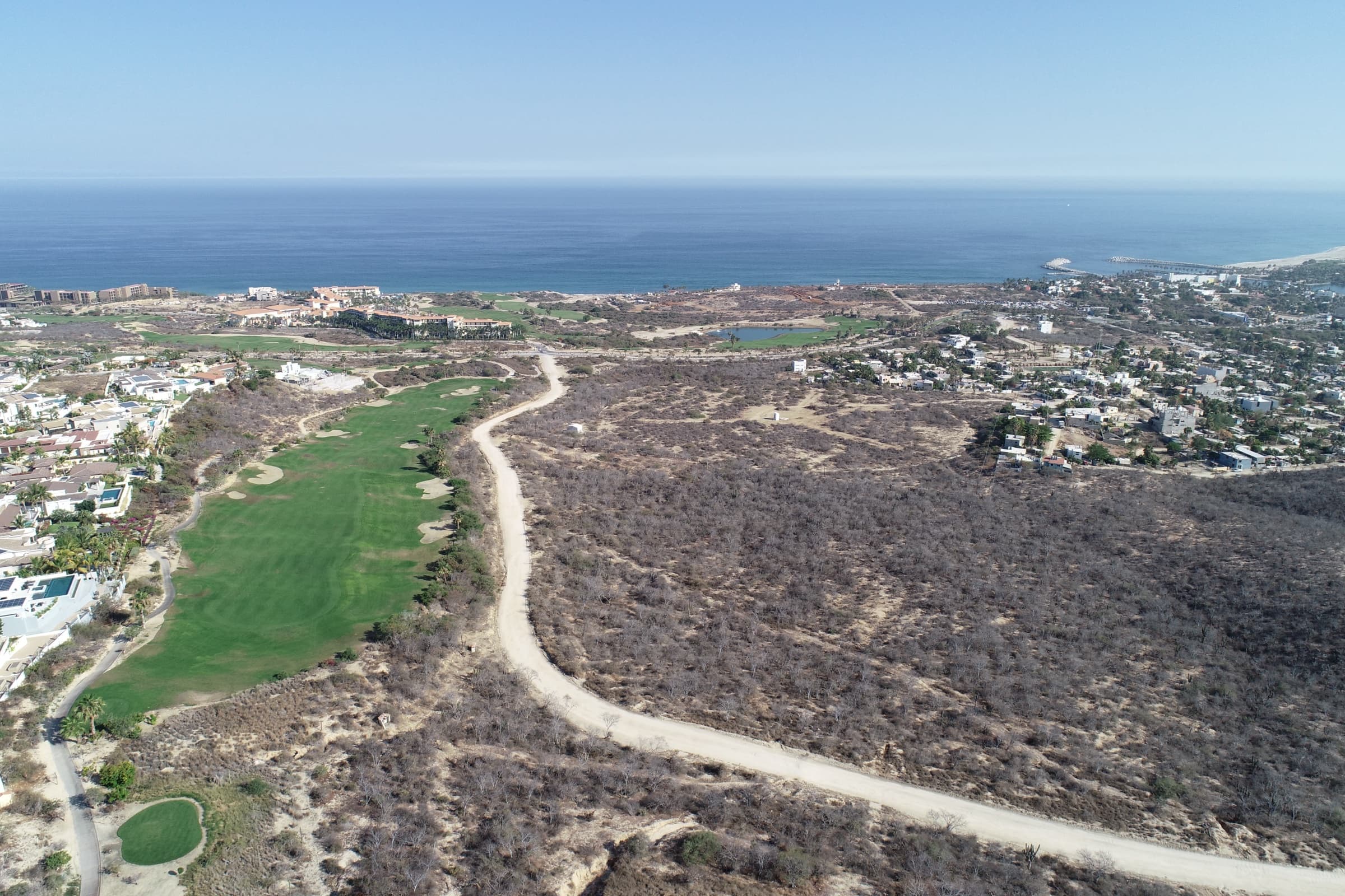 Elevated panoramic aerial of the Vitality Grove site with golf course fairway, coastline, ocean horizon, and distant mountains.